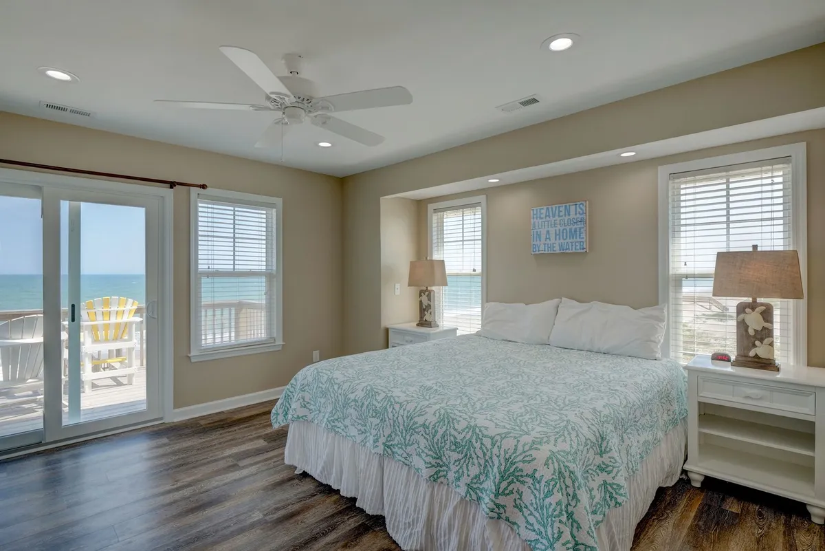 A serene king bedroom with coral-patterned bedding is filled with natural light from large windows that offer an expansive ocean view. The room features wooden flooring and tasteful decorative lamps, creating a tranquil and inviting ambiance ideal for rest and relaxation.