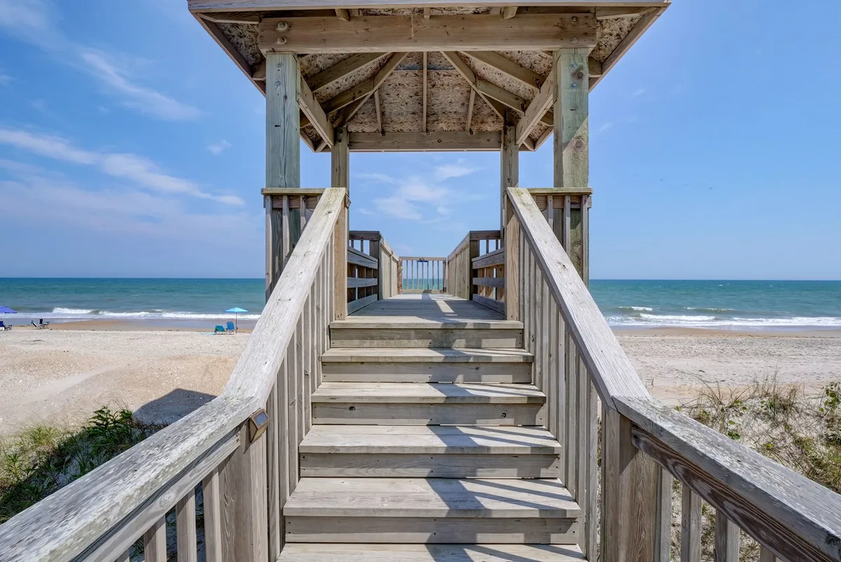 A set of wooden stairs leads down toward a peaceful beach and ocean horizon, with a small gazebo structure at the edge. The scene is serene and open, inviting walks to the shore for refreshing breaks. The sky and water create a calming backdrop, reinforcing the restorative qualities of the setting.