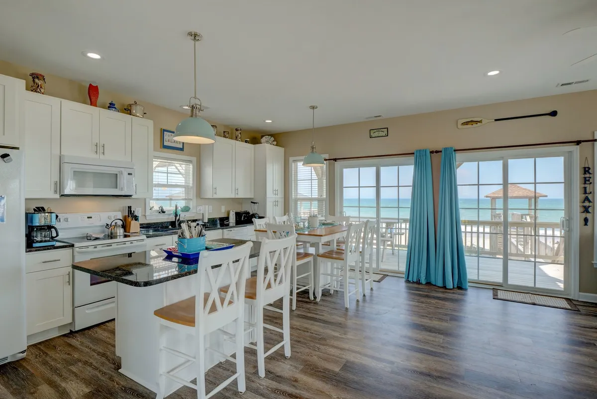 This image features a welcoming, open-concept kitchen and dining area with a kitchen island that offers seating, large windows framing an expansive ocean view, and a dining table with chairs. The setting is bright, modern, and casual, inviting guests to enjoy meals, morning coffee, or evening gatherings with the sea as a backdrop.