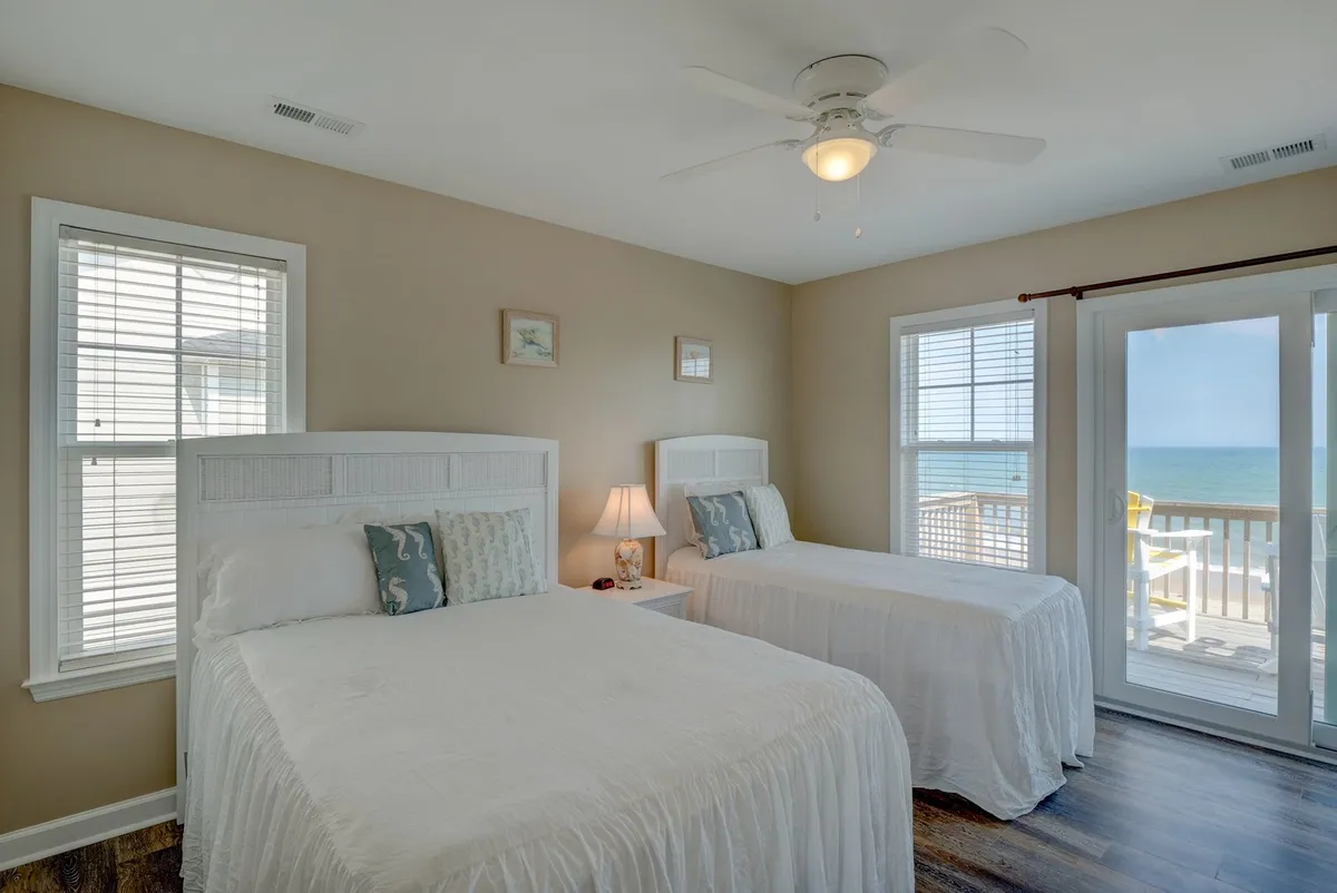 A serene bedroom features two beds with crisp white linens. Through a sliding glass door, the ocean and part of the deck are visible, suggesting the ease of stepping out from the bedroom directly onto a private deck. The tranquil mood hints at the possibility of enjoying the sunrise from bed or the deck.