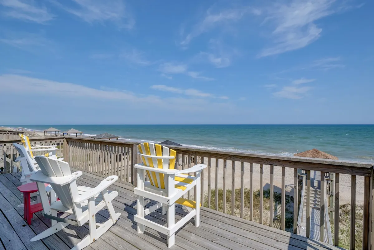 An expansive oceanfront deck is furnished with classic Adirondack chairs positioned to face a sweeping view of the sea. A wooden walkway leads directly from the deck toward the sandy beach below. The scene is bathed in soft natural light, emphasizing the serene ambiance ideal for relaxation, reading, or sipping morning coffee while watching the sunrise.
