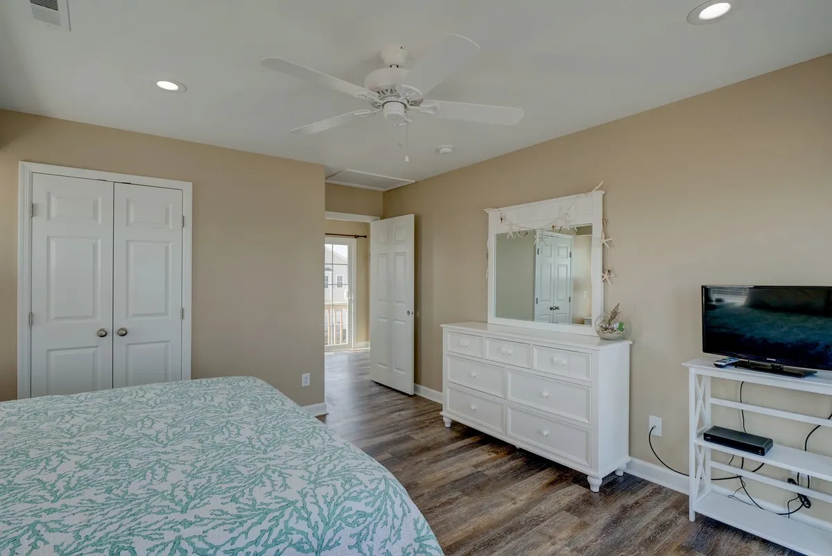 A cozy bedroom featuring a bed with stylish patterned bedding, a white dresser, a mirror, and a television. Natural light streams in, adding to the calm, serene mood—ideal for peaceful mornings or relaxing evenings.