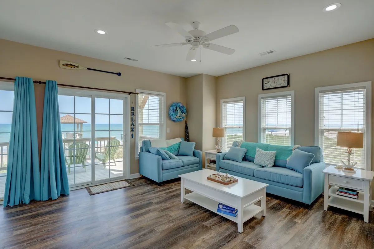 The living room features plush light blue sofas, a white coffee table, large windows offering an unobstructed view of the ocean, and coastal-inspired decorative elements. The space exudes calm and is well-suited for relaxation, socializing, or enjoying cable TV.