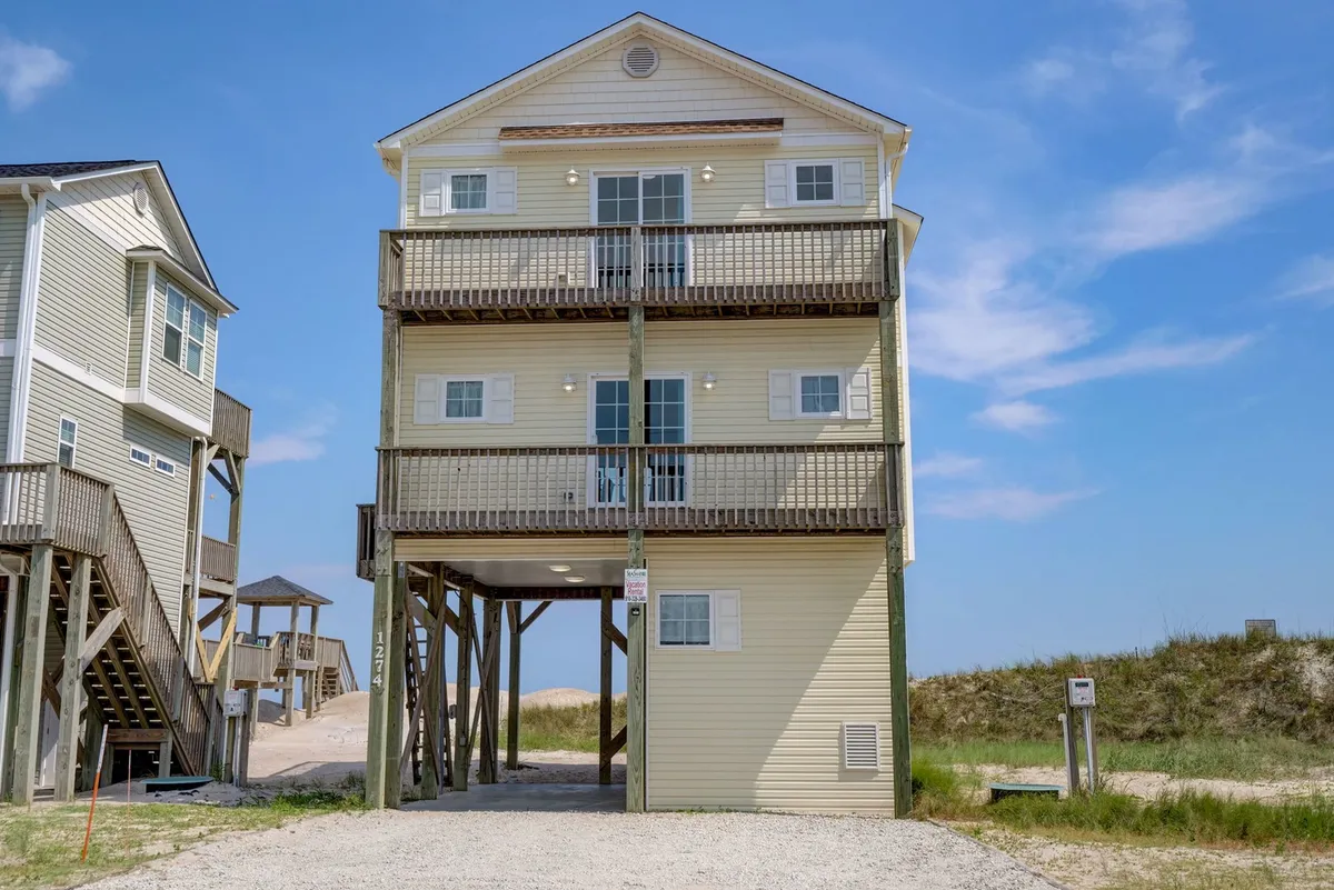 A cheerful view of a three-story oceanfront house supported by wooden beams, with multiple balconies facing the water. The scene suggests ample outdoor space to enjoy the sights and sounds of the ocean, and the structure’s orientation hints at striking sunrise and sunset views from the decks.