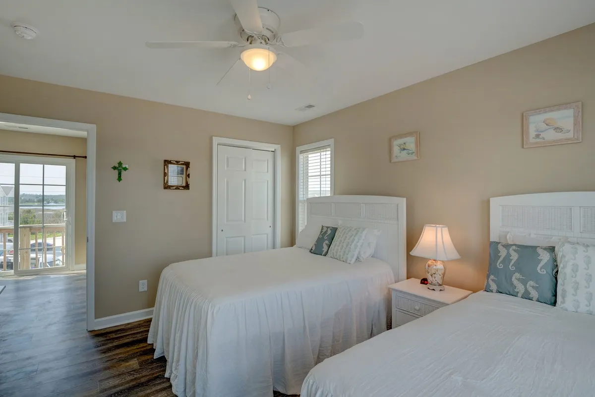 A serene bedroom with two twin beds, a window letting in natural light, a ceiling fan, and a decorative lamp on the nightstand. The layout feels peaceful and practical, suggesting restful nights and family-friendly accommodations.