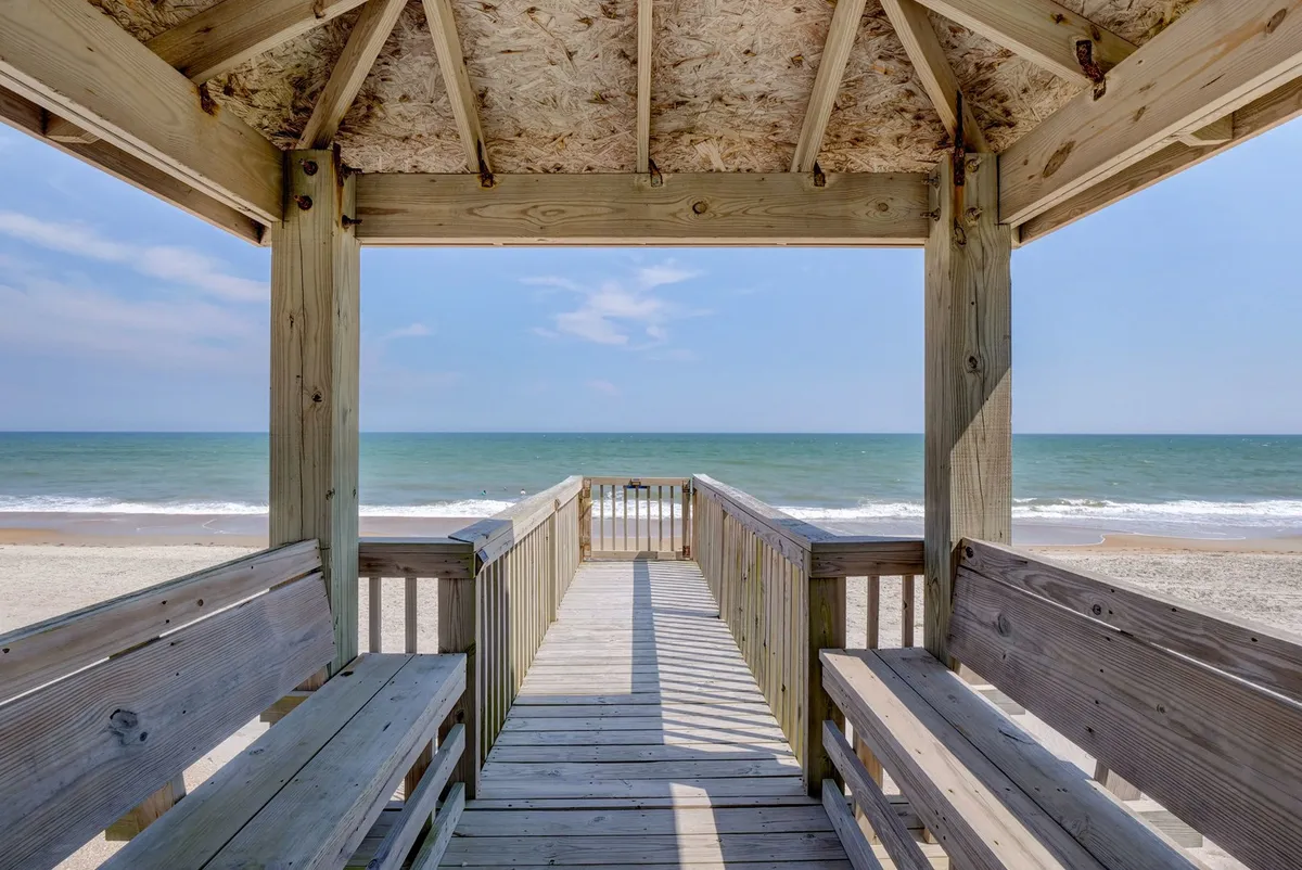 An inviting wooden walkway leads over sandy dunes toward the open ocean, with a vast expanse of sky and gentle waves in the distance. The scene is calm and serene, evoking a sense of peaceful escape and direct beach access for quiet walks or intimate moments.