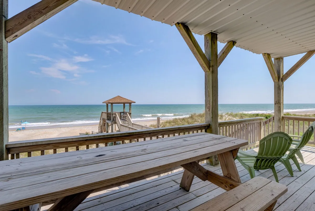 A sun-drenched outdoor seating area on a wooden deck, with a clear view of the ocean. There's a table and chairs set for outdoor dining or focused work, and a playground structure is visible, suggesting options for family-friendly breaks. The mood is casual and welcoming, emphasizing relaxation and enjoyment of the surroundings.