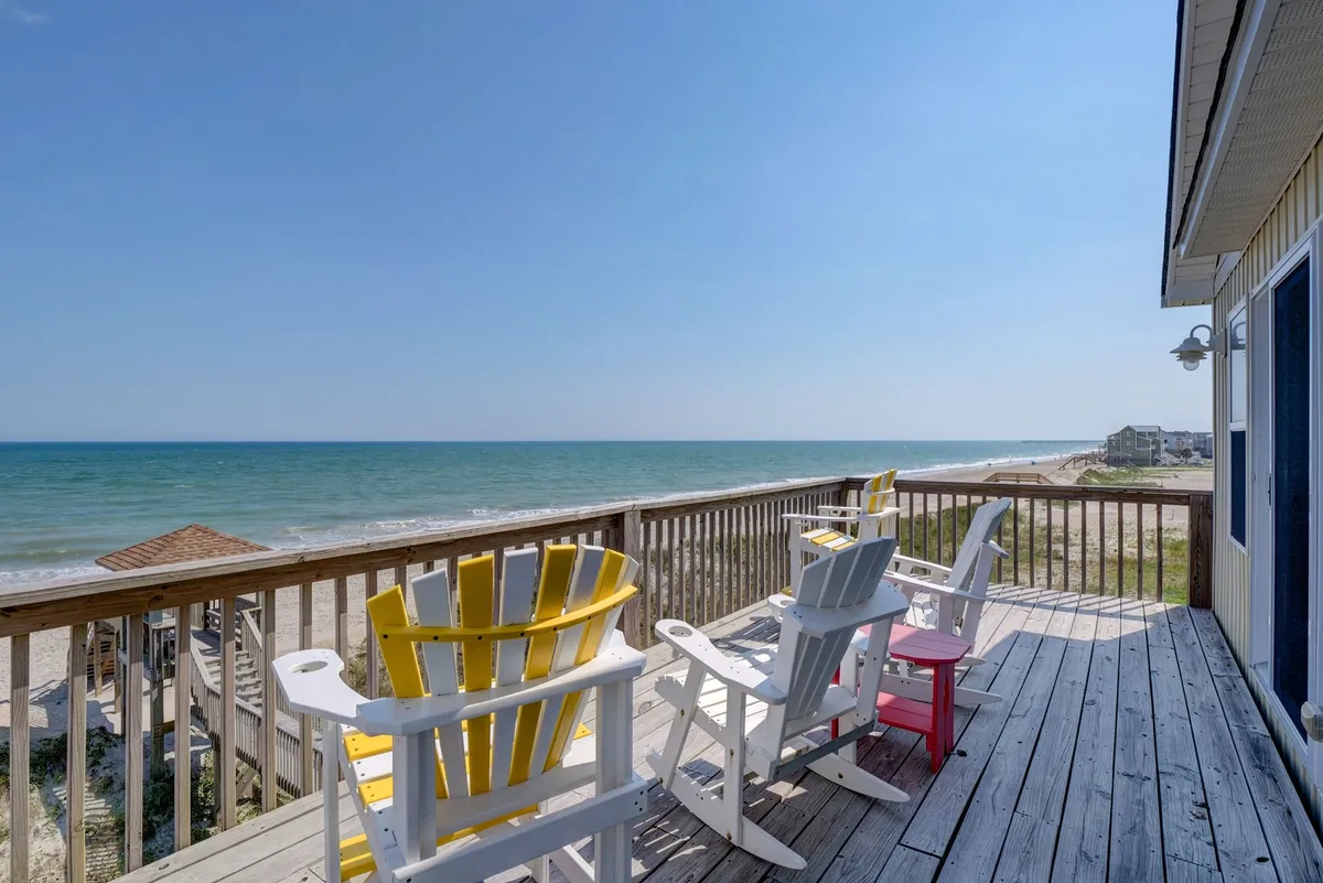 The image showcases a spacious wooden deck overlooking the Atlantic Ocean, featuring a row of colorful Adirondack chairs perfectly arranged to face the sparkling water. The scene is bathed in natural light, inviting relaxation, while the sweeping ocean view promises tranquil mornings and breathtaking sunsets. The atmosphere feels welcoming and laid-back, ideal for unwinding, sharing moments with loved ones, or simply soaking in the beauty of North Topsail Beach.