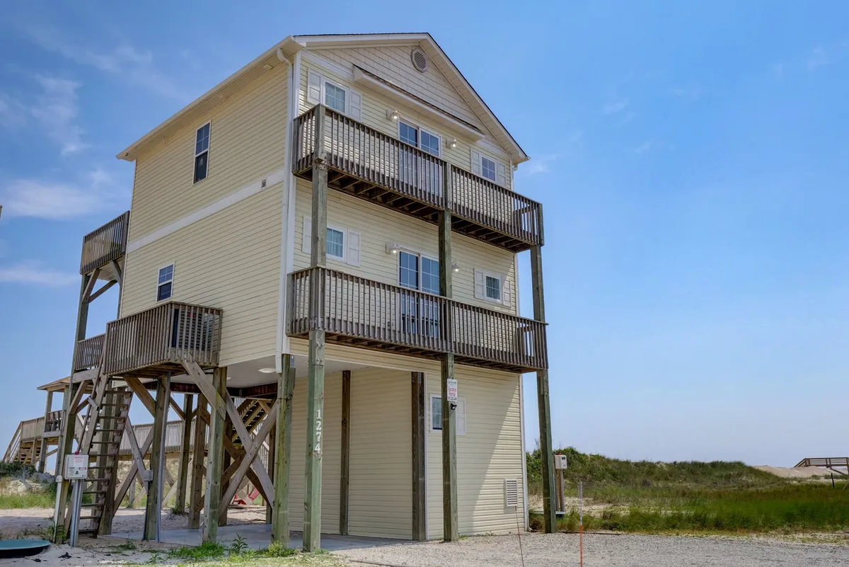 A three-story beachfront house elevated on stilts, featuring broad balconies that overlook the ocean. The cheerful exterior and ample balcony spaces are designed for relaxation and outdoor living, showcasing the property's proximity to the ocean and suitability for enjoying the sea breeze while working or unwinding.