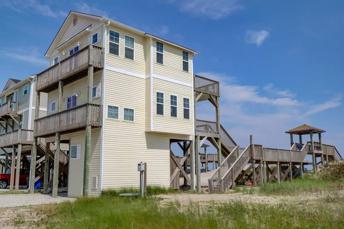 A multi-level beach house features spacious wooden decks at different elevations, connected by a set of stairs that leads straight down to the beach. Surrounding grassy areas and open sky frame the structure, suggesting ample space for outdoor dining, sunbathing, or gathering with friends and family.