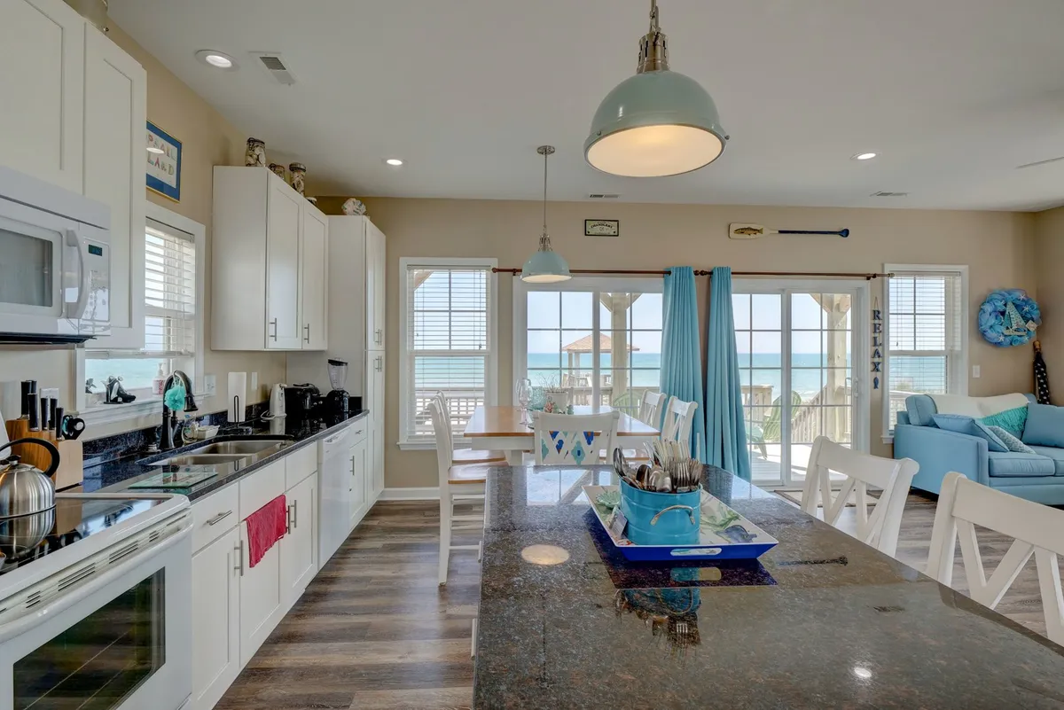 A modern kitchen scene showcasing a spacious island with a dark granite countertop, blue accents in the decor, and large windows that overlook the ocean. The natural light and layout emphasize a casual, cheerful setting ideal for both entertaining and relaxed family time.