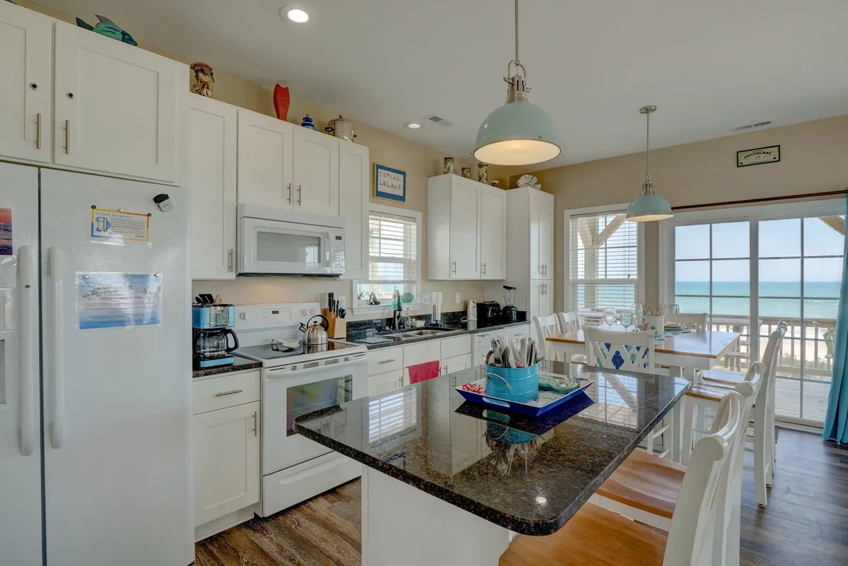 A bright, cheerful kitchen with a large island and a window framing ocean views, perfect for gathering the family for meals after a day on the beach. Decorative elements and relaxed seating foster a welcoming, communal atmosphere.